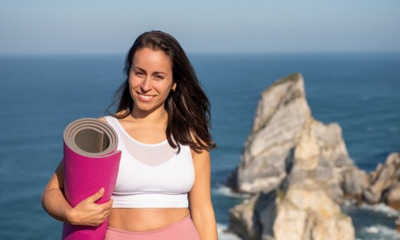 Smiling woman with yoga mat in sportswear by oceanic cliffs in Portugal.